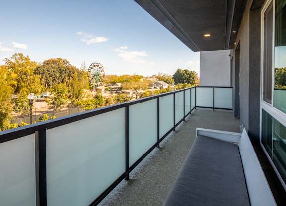 A balcony with a glass railing and a bench overlooks a park with a Ferris wheel.