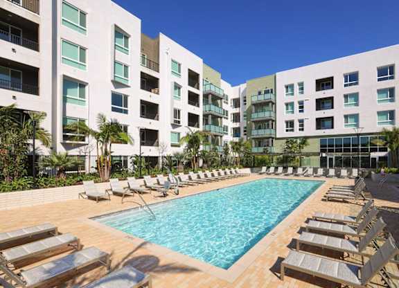A swimming pool surrounded by sun loungers in front of apartment buildings.