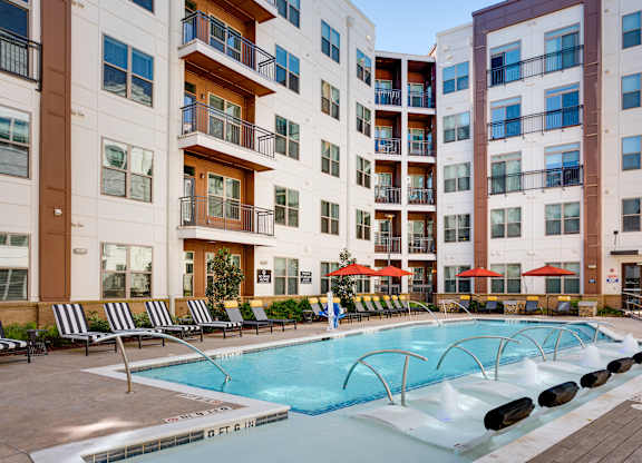 a swimming pool with chaise lounge chairs and umbrellas in front of an apartment building
