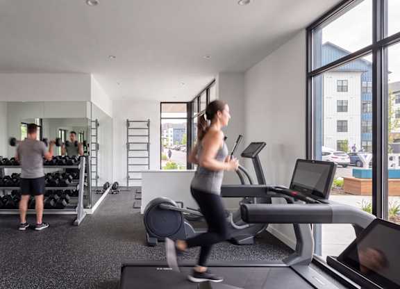 A woman is running on a treadmill in a gym with a man standing behind her.