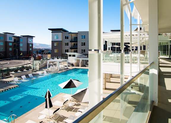 A large swimming pool with sun loungers and umbrellas is surrounded by apartment buildings.