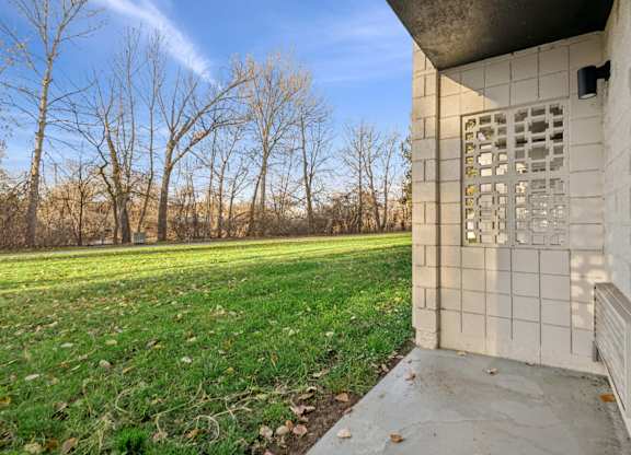 A concrete building with a window and a door is in the foreground with a grassy field and trees in the background.