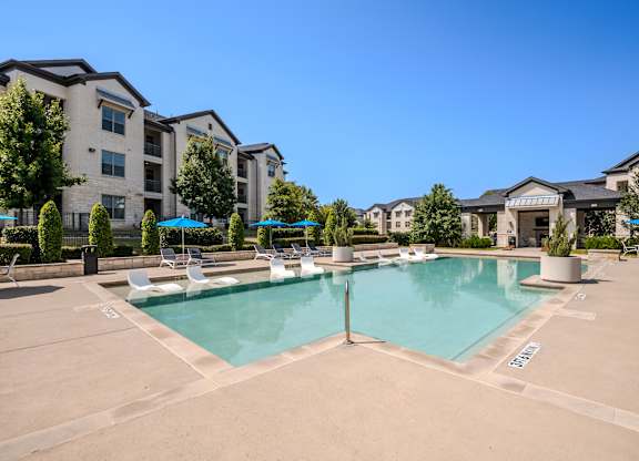 A large swimming pool surrounded by lounge chairs and umbrellas in front of apartment buildings.