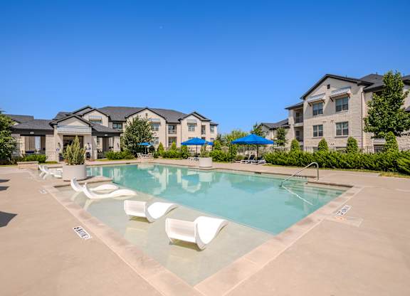 A swimming pool surrounded by white lounge chairs and apartment buildings in the background.