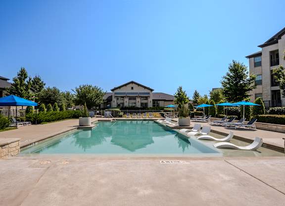 A large swimming pool with a stone border and a stone patio with lounge chairs and umbrellas.