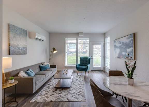 A living room with a grey sofa, a coffee table, a dining table, and a large window.