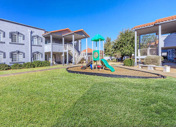 A playground with a green slide and a blue umbrella is in front of a building.