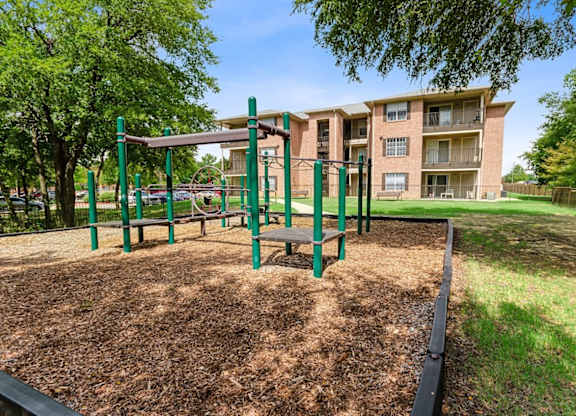 A playground with a green swing set and a brown mulched area in front of a brick apartment building.