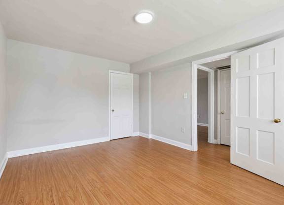 A room with white walls and wooden flooring, with two doors and one open to a hallway.