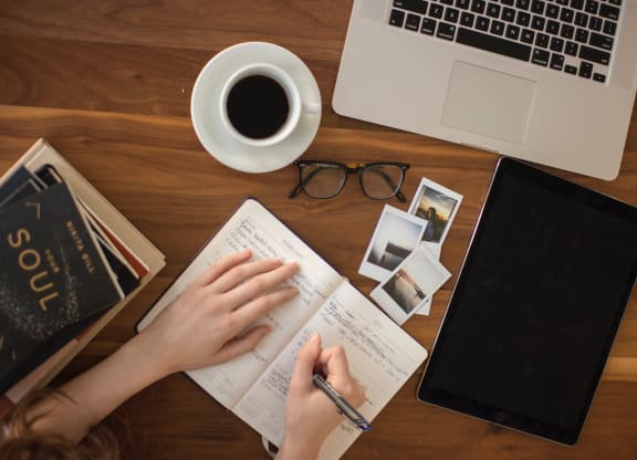 person writing in journal with laptop, notebook and cup of cofee