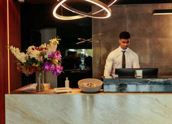 A man in a white shirt and tie is standing behind a reception desk with a laptop and a bowl of candy.