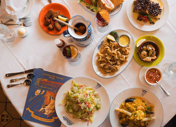 A table with a white tablecloth is set with plates of food, glasses, and a menu.