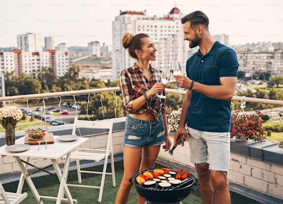 A man and woman are standing on a balcony with a barbecue grill in front of them.