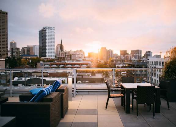 A rooftop patio with a table and chairs overlooking a city skyline at sunset.