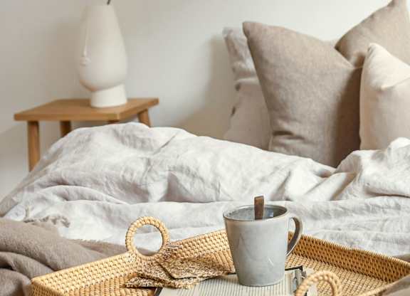A grey mug with a wooden handle sits on a woven tray with a white lamp and pillows in the background.