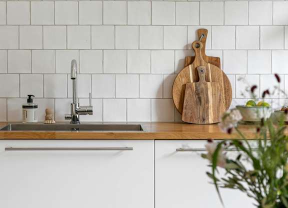 A kitchen with a white tile backsplash and a wooden cutting board on the counter.