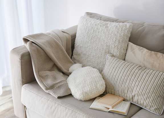 A beige couch with a white pillow, a white knitted hat, a book and a grey blanket.