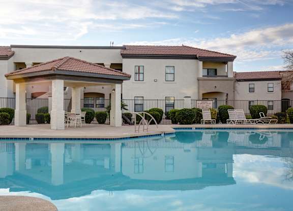 A swimming pool in front of a building with a patio and chairs.