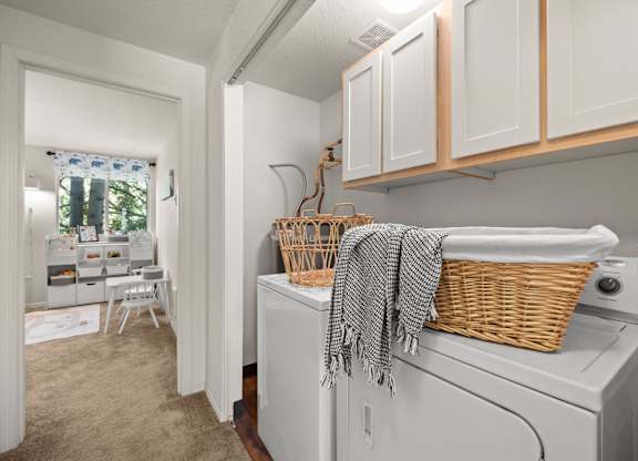 A laundry room with a washer and dryer, a basket on top of the dryer, and a window with a view of trees at Clackamas Trails Apartments, Oregon, 97222
