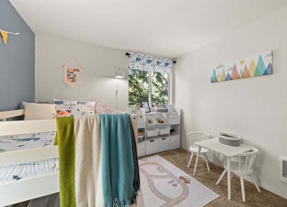 A white bed with a green, blue, and white blankets is in a room with a white table and chairs at Clackamas Trails Apartments, Oregon, 97222