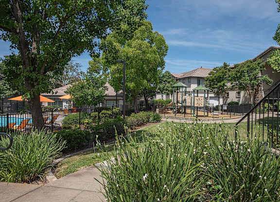A view of a swimming pool gate and playground with trees at Stoneridge Apartments, California, 95661