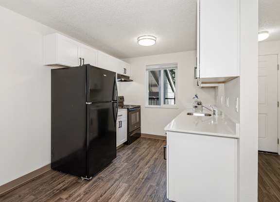 A black refrigerator stands in a kitchen with white cabinets and a white sink at Todd Village Apartments, Oregon, 97062