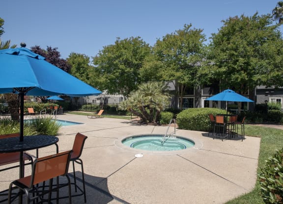 outdoor spa with manicured landscaping and patio tables with orange chairs and blue umbrellas at Woodbridge Apartments, California