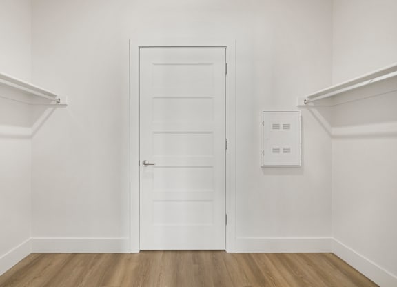 A white door is in the middle of a room with white walls and wood flooring.