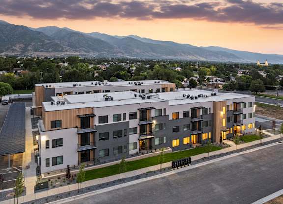 A modern apartment building with a parking lot in front and mountains in the background.