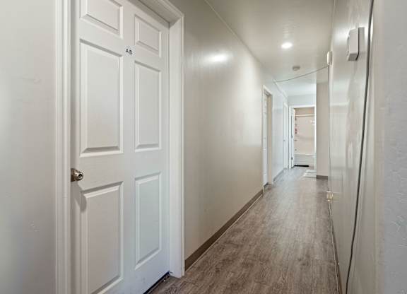 A white door with a gold handle is on the left of a hallway with white walls and wood flooring.