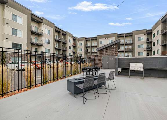 A patio with a table and chairs is surrounded by a black fence and apartment buildings.
