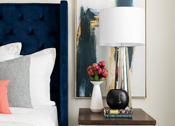 A bedroom with a bed, a side table with a lamp and a painting on the wall.