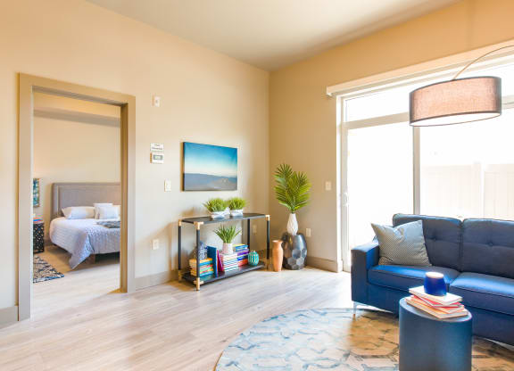 a living room with a blue couch and a lamp and a bedroom in the background at Sugar House Apartments By Urbana, Salt Lake City, UT, 84105