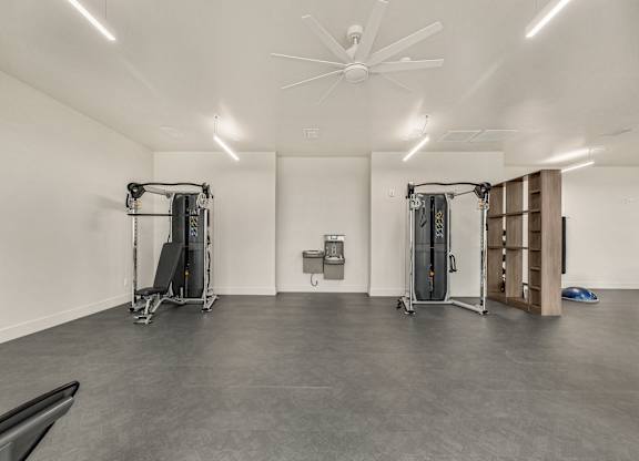 A gym with a grey floor and white walls has a fan and two pieces of gym equipment.