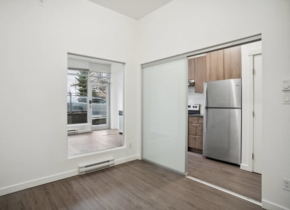 an empty room with a door to a kitchen and a refrigerator