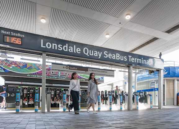 Two women walking in a station with a sign that says Lonsdale Quay SeaBus.