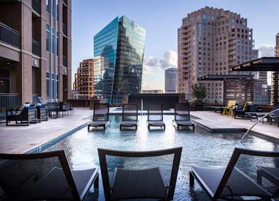 A pool with chairs around it in the foreground with buildings in the background.