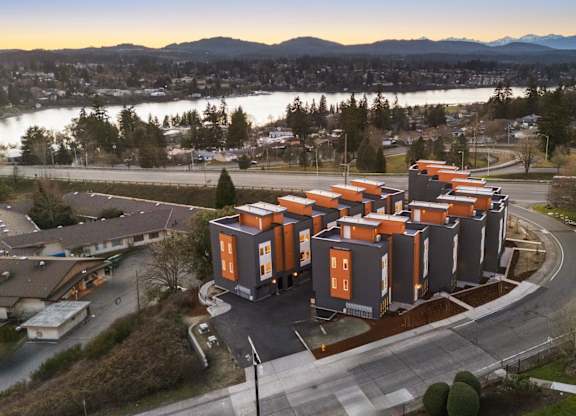 A modern building with orange and grey exterior is surrounded by a parking lot and a body of water in the background. at The Narrows at Clare Apartments , Bremerton, 98310