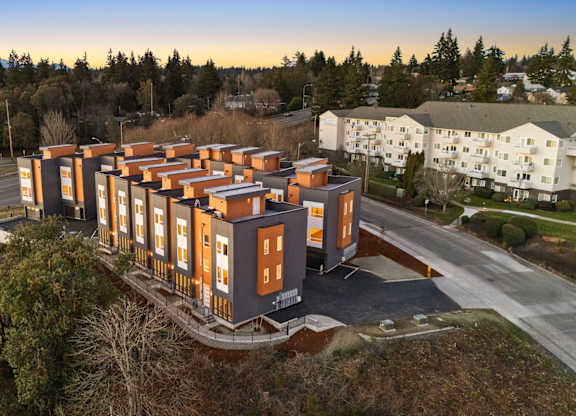 A modern apartment complex with orange and white buildings is surrounded by greenery and a road. at The Narrows at Clare Apartments , Apartments, Bremerton, WA