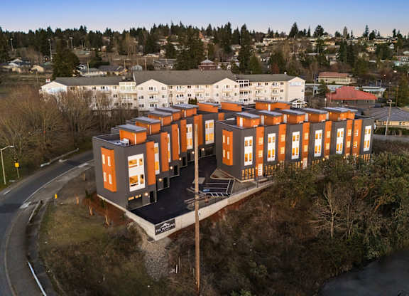 A modern building with orange and white exterior is situated on a small hill with a road and other buildings in the background. at The Narrows at Clare Apartments , Bremerton, 98310