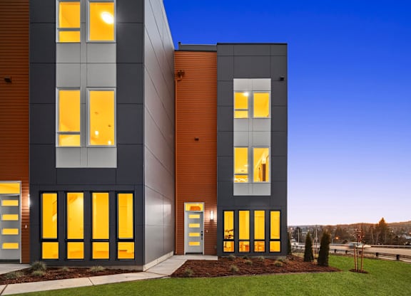 A modern building with a combination of grey and brown exterior and yellow lit windows at The Narrows at Clare Apartments , Washington, 98310
