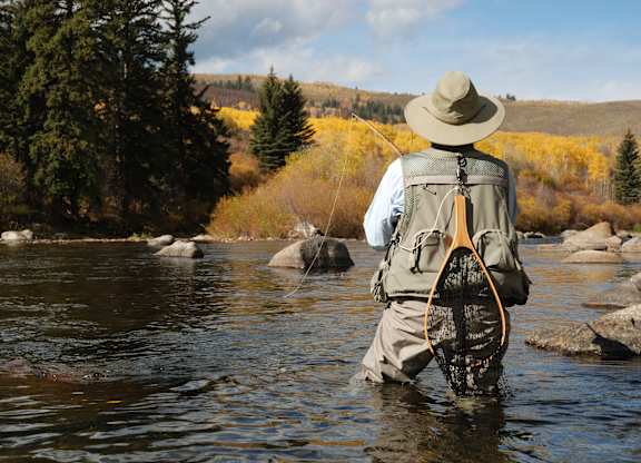 A man in a hat and waders stands in a river holding a fly fishing net.