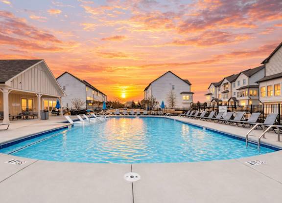 A swimming pool surrounded by lounge chairs and buildings under a sunset sky.