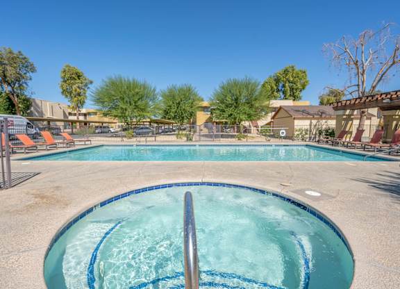 A swimming pool and a hot tub that is gated and surrounded by lounge chairs.