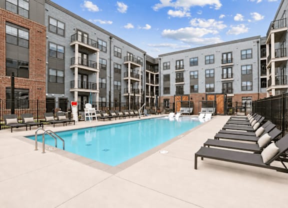 A swimming pool is surrounded by lounge chairs in front of apartment buildings.