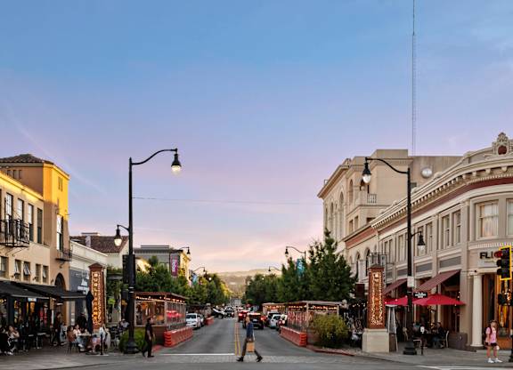 A street view of a city with buildings on either side and people sitting at tables outside the restaurants.