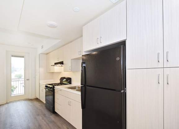 A black refrigerator stands in a kitchen with white cabinets and a wooden floor.