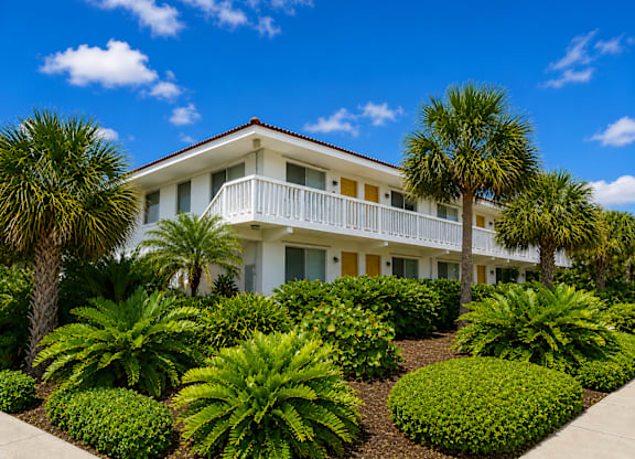799 - A white building with a balcony is surrounded by green plants.
