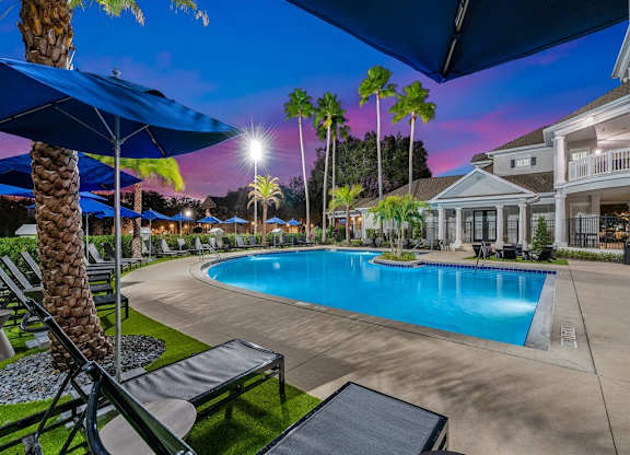 our resort style swimming pool is lit up at night with palm trees