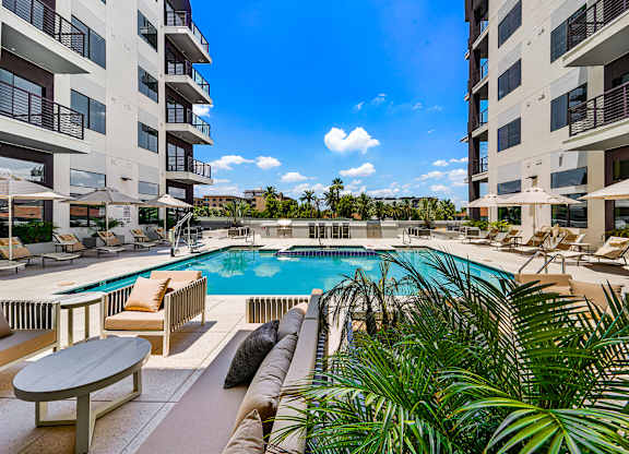 A pool surrounded by chairs and plants with apartment buildings in the background.